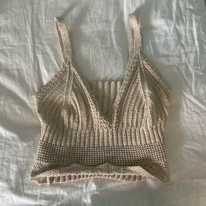 Beige stickad linnetopp - Säljer en superfin beige stickad linnetopp. Den har en v-ringad design och är croppad, vilket gör den perfekt för sommardagar. Toppen är ärmlös med breda axelband och har en ribbad strukt. Den är virkad och är guld glittrig, köpte på loppis, som ny