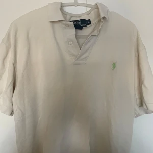 Beige pikétröja från Ralph Lauren - Säljer en klassisk beige pikétröja från Ralph Lauren i storlek M. Tröjan har en stilren design med en liten grön broderad logga på bröstet. Perfekt för en avslappnad men ändå snygg look. Den har korta ärmar och knappar vid kragen.