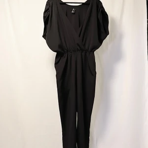 Svart jumpsuit med omlott - Snygg svart jumpsuit med omlott-design och kortärmade puffärmar. Knappt använd. Perfekt för en festlig kväll eller en stilren vardagslook. Den har en elastisk midja för en bekväm passform. Rymlig & lite oversize. 