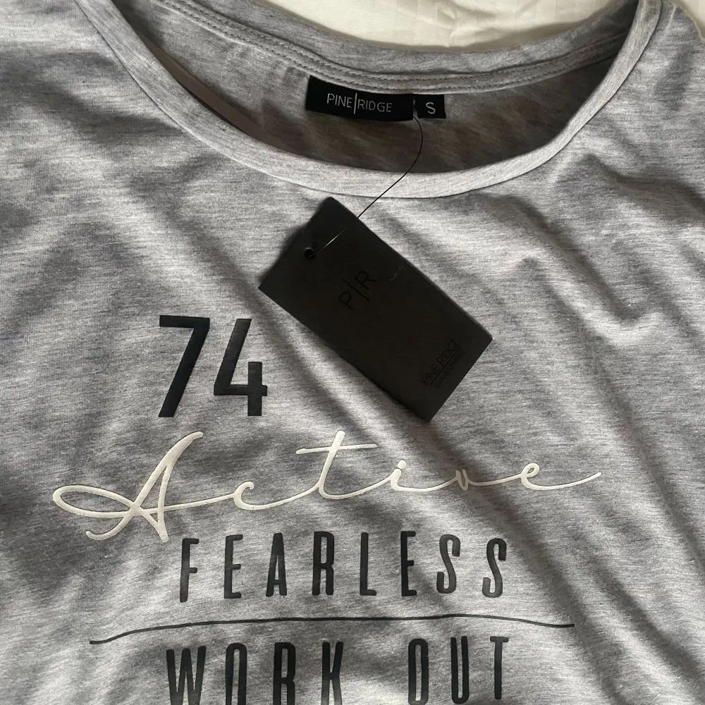 Ny grå träning tröja från Pine Ridge i storlek S. I ett super mjukt material. Tryck med texten '74 Active Fearless Work Out' på framsidan. Tröjan är kortärmad och har en rund halsringning.. T-shirts.