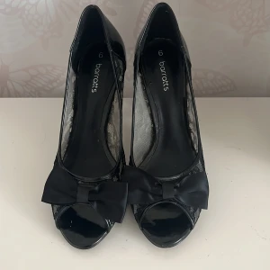 Svarta klackskor med spets och rosett - Säljer ett par eleganta svarta klackskor från Barratts. De har en snygg spetsdesign på sidorna och en fin rosett framtill. Skorna har en öppen tå och en smal klack som ger en stilren look. Perfekta för festliga tillfällen!