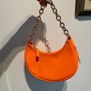 Orange axelväska med kedjedetaljer - Säljer en supercool orange axelväska med en chunky kedja som axelrem. Väskan har en dragkedja upptill och är perfekt för att liva upp vilken outfit som helst. Den är i syntetmaterial och har en stilren design som passar både till vardags och fest.
