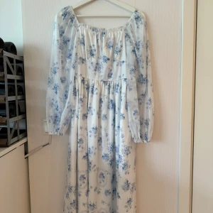 Vit och blå blommig långklänning - Säljer en superfin vit långklänning med blått blommönster. Klänningen har långa ärmar och en luftig passform, perfekt för sommarens fester eller en mysig vårdag. Den är gjord i ett lätt och bekvämt material som känns skönt mot huden.