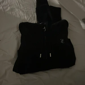 Svart hoodie från Juicy Couture - Säljer en svart hoodie från Juicy Couture med dragkedja och huva. Den har långa ärmar och texten 'Juicy' i glittriga bokstäver på ryggen. Perfekt för en avslappnad och stilren look!