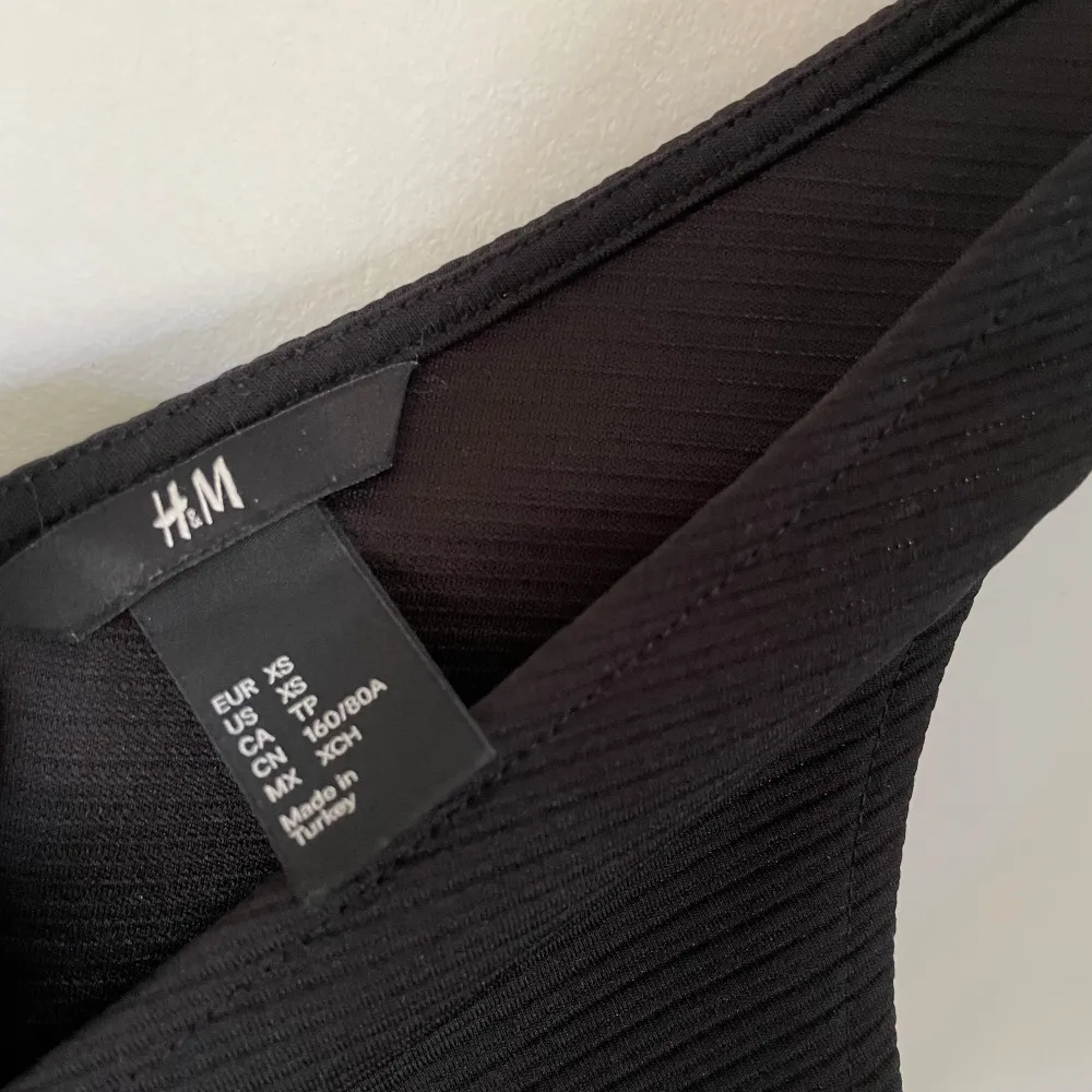 Säljer en elegant svart klänning från H&M i storlek XS. Klänningen har en v-ringad design och är ärmlös, vilket gör den perfekt för både fest och mer formella tillfällen. Den har en dragkedja på sidan för enkel påklädning och en stilren, figursydd. . Klänningar.