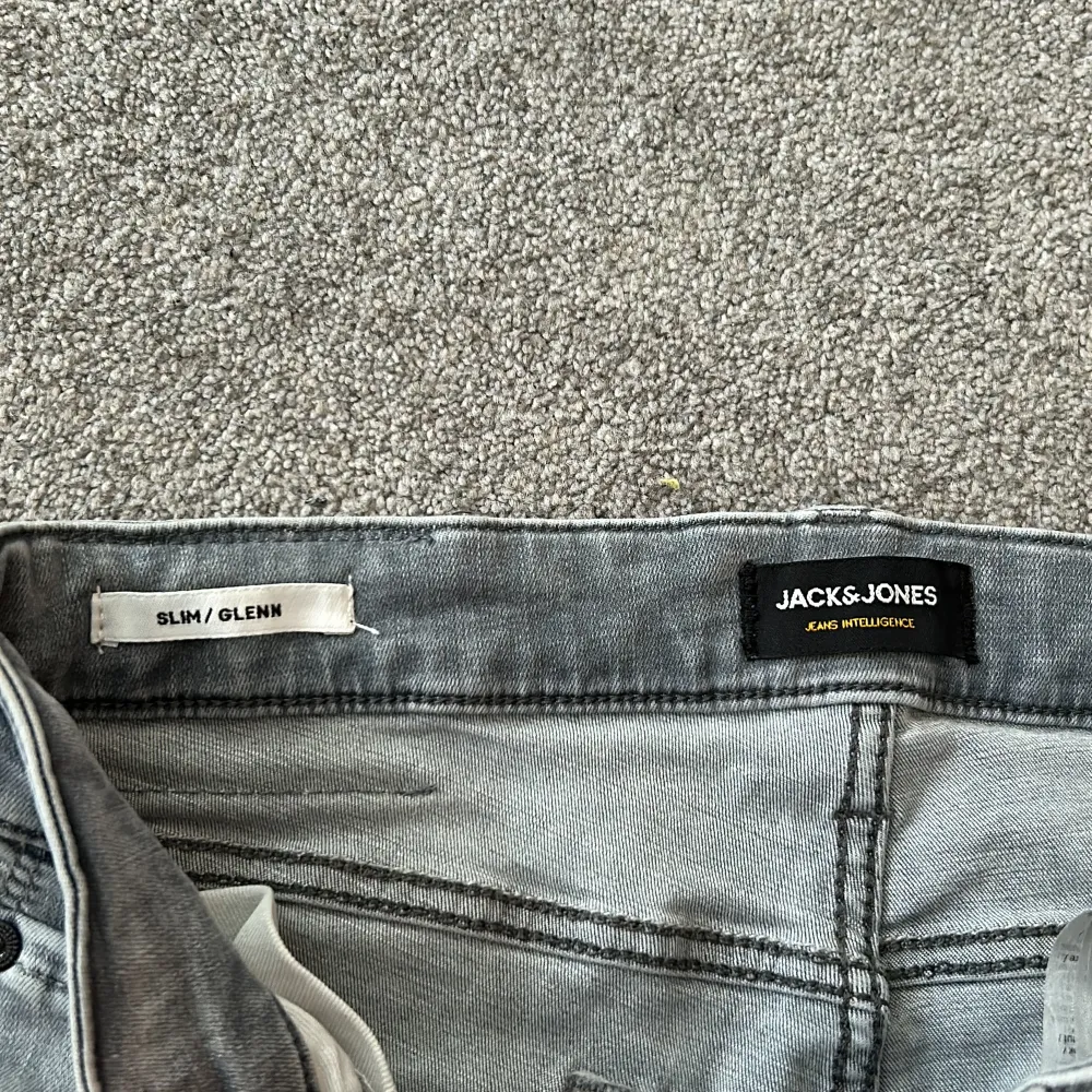 Säljer ett par snygga grå jeans från Jack & Jones i modellen Slim/Glenn. De har en slim passform och är perfekta för en stilren look. Jeansen är i bra skick och har en klassisk femficksdesign med en läderpatch på baksidan. Perfekta för både vardag och fest!. Jeans & Byxor.