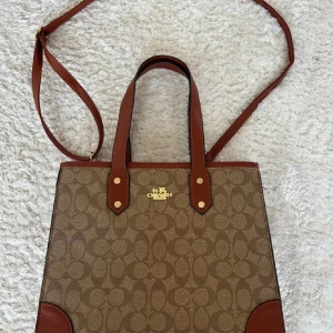 Brun axelväska frän Coach - Saljer en snygg brun axelvaska. Vaskan har ett klassiskt mönster med Coach-loggan, osäker pa äkthet dock. Den har bäde handtag och en justerbar axelrem for flexibel anvandning. Vaskan har en dragkedja upptill och flera fack inuti för enkel organiseri