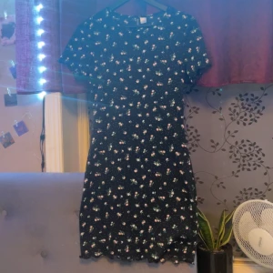 Svart blommig klänning - Säljer en söt svart klänning med blommigt mönster i rosa och grönt. Den har korta ärmar och en bekväm passform som passar perfekt för sommarens alla tillfällen. Klänningen är i bra skick och har en fin volangdetalj längst ner och även vid armarna. 