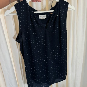 Svart ärmlös blus från Holly & White - Snygg marinblå ärmlös blus från Holly & White med små vita prickar. Perfekt för både vardag och fest. Blusen har en lös passform och är gjord i ett lätt och bekvämt material. Passar bra till både jeans och kjol!