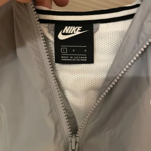 Grå och vit vindjacka från Nike - Säljer en snygg grå och vit vindjacka från Nike i storlek L. Jackan har en dragkedja framtill och en huva för extra skydd mot väder. Perfekt för vår och höst! 🌀