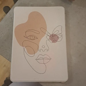 iPad-fodral med konstnärligt ansikte - Säljer ett stilrent iPad-fodral med ett konstnärligt motiv av ett ansikte i linjeteckning och en fjäril. Fodralet är i ljusa toner av beige och rosa, vilket ger en modern och elegant look. Perfekt för att skydda din iPad med stil! 💼✨