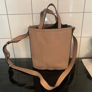 Beige axelväska i skinn - Säljer en snygg beige axelväska i skinn med justerbar axelrem och praktiska handtag. Väskan har en stilren design och stängs med en knapp. Perfekt för vardagsbruk och rymmer det mesta du behöver ha med dig. Inuti finns ett fack med dragkedja för extra säkerhet.