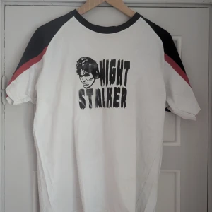 Vit t-shirt med tryck 'Night Stalker' - Cool vit t-shirt med svart tryck 'Night Stalker' och en illustration på framsidan. T-shirten har korta ärmar med svarta och röda detaljer på ärmarna. Perfekt för en unik och edgy stil.