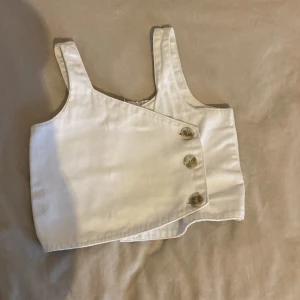 Vit omlott topp - Säljer en stilren vit omlott topp med breda axelband och knappar framtill. Toppen har en elastisk del i ryggen för bättre passform. Perfekt för en somrig look! 💕