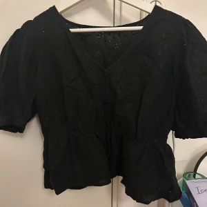 Svart blus från LCW Casual - Säljer en svart blus från LCW Casual med korta puffärmar och v-ringning. Blusen har knappar framtill och ett diskret broderat mönster. Perfekt för en stilren look.