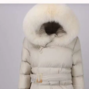 Beige dunjacka med pälsdetalj - Säljer en elegant beige dunjacka med stor huva kantad av fluffig päls. Jackan har ett bälte i midjan för en snygg passform och är perfekt för kalla dagar. Den är både stilren och funktionell, vilket gör den till ett måste i vintergarderoben.
