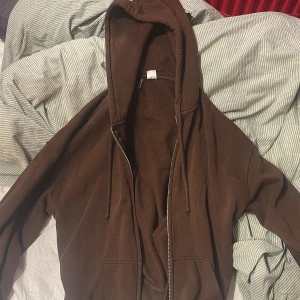 Brun hoodie med dragkedja - Säljer en brun hoodie med dragkedja och huva. Perfekt för kyliga dagar med sin mysiga känsla. Den har långa ärmar och snören vid huvan för justering. Enkelt och stilrent plagg för vardagsbruk.
