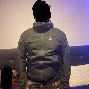 Haglöfs jacka - Säljer en svart vindjacka från Haglöfs med huva och dragkedja framtill. Jackan har en enkel och stilren design, perfekt för blåsiga dagar. Den är långärmad och har justerbar snörning i huvan för extra skydd. Storlek M men