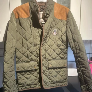 Grön quiltad jacka från Lexington - Säljer en snygg grön quiltad jacka från Lexington med bruna axelpartier i mocka. Jackan har broderad logga på bröstet och knappar framtill. Perfekt för höst och vår!