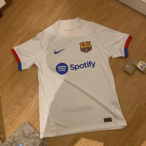 Vit FC Barcelona fotbollströja från Nike - Säljer en vit FC Barcelona fotbollströja från Nike med korta ärmar. Tröjan har klubbens emblem och sponsorloggor i blått och rött. Tillverkad i Dri-FIT material för optimal komfort. Perfekt för fans som vill visa sitt stöd. Inte använd.