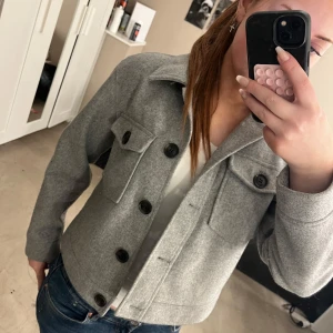 Grå kavaj med stora knappar - Snygg grå kavaj med stora svarta knappar och två fickor framtill. Jackan har en klassisk krage och är i en croppad modell som ger en modern look. Perfekt för att lyfta vilken outfit som helst!