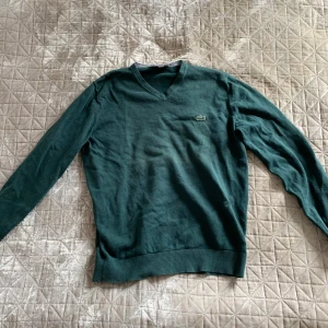 Grön tröja från Lacoste - Säljer en stilren grön tröja från Lacoste med v-ringning och det klassiska krokodilmärket på bröstet. Tröjan har långa ärmar och ribbade muddar vid ärmslut och nederkant. Perfekt för en avslappnad och snygg look.