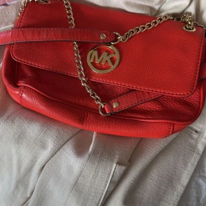 Röd axelväska från Michael Kors - Snygg röd axelväska från Michael Kors med kedjedetaljer och logotyp i guld. Väskan har ett rymligt fack med dragkedja och ett stilrent foder med Michael Kors-mönster. Perfekt för att ge en färgklick till din outfit! Den har lite skador som ni ser på bilderna!!