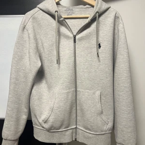 Grå hoodie från Polo Ralph Lauren - Säljer en stilren grå hoodie från Polo Ralph Lauren med dragkedja och broderad logga på bröstet. Tröjan har en klassisk passform med långa ärmar och justerbar huva. Perfekt för en avslappnad look.