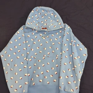 Blå hoodie med Hello Kitty-mönster från Nike - Säljer en blå hoodie från Nike med ett gulligt Hello Kitty-mönster över hela tröjan. Hoodien har en klassisk passform med en stor ficka framtill och en justerbar huva. Perfekt för dig som älskar unika och lekfulla plagg!