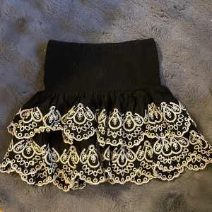 Svart kjol med broderade detaljer - Snygg svart kortkjol med vackra vita broderade detaljer och volanger. Perfekt för att ge en unik touch till din outfit. Den har en elastisk midja för bekväm passform.