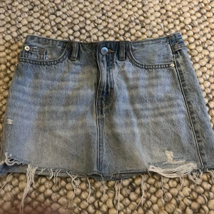 Blå jeanskjol med slitningar - Snygg blå jeanskjol med slitningar och fransig kant. Kjolen har en klassisk femficksdesign och en knapp framtill. Perfekt för en avslappnad stil.