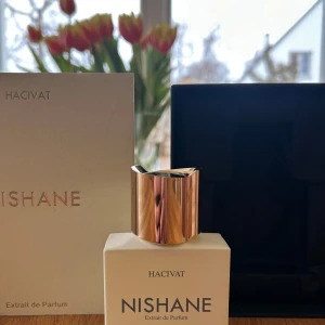 Hacivat Extrait de Parfum från Nishane - Säljer en exklusiv Hacivat Extrait de Parfum från Nishane. Flaskan har en elegant design med en vit kropp och en glänsande guldfärgad kork. Parfymen kommer i en stilren svart och vit förpackning. Perfekt för den som söker en lyxig doftupplevelse.