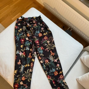Blommiga byxor från Stradivarius - Snygga blommiga byxor från Stradivarius med ett färgglatt mönster på svart bakgrund. Byxorna har en elastisk midja och är perfekta för en somrig look. De är tillverkade i ett lätt och bekvämt material.
