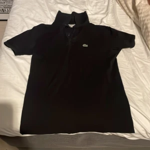 Svart pikétröja från Lacoste - Säljer en klassisk svart pikétröja från Lacoste. Tröjan har korta ärmar och den ikoniska krokodilloggan på bröstet. Perfekt för en stilren och avslappnad look. Passar både till vardags och mer uppklädda tillfällen.
