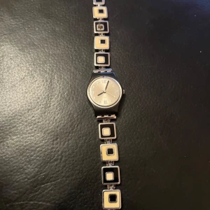 Svart och silver klocka från Swatch - Snygg klocka från Swatch med svart urtavla och silverdetaljer. Armbandet har ett unikt mönster med fyrkantiga länkar i svart och silver. Klockan är vattentålig och tillverkad i Schweiz. Perfekt för dig som gillar stilrena accessoarer.