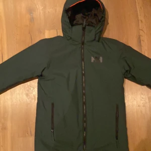 Grön skidjacka från Helly Hansen - Säljer en grön skidjacka från Helly Hansen med huva och dragkedja. Jackan är perfekt för kyligare väder och har en stilren design med Helly Hansen-logga på bröstet. Den är tillverkad i slitstarkt material och har praktiska fickor med dragkedjor. Storlek 164 eller 14 år. Använd endast 1 gång ord pris 1100kr