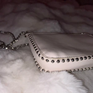 Cream vit axelväska med kedja - Snygg vit axelväska med silverkedja och nitar längs kanterna. Väskan har en dragkedja upptill för säker förvaring. Perfekt för att ge en edgy touch till din outfit.