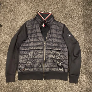Svart jacka från Tommy Hilfiger - Marinblå cardigan från Tommy Hilfiger med quiltad framsida och dragkedja. Jackan har långa ärmar och en ribbad krage med röd och vit detalj. Perfekt för en stilren look. Ny pris runt 2500kr
