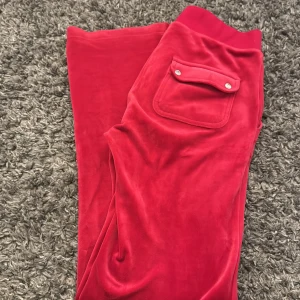 Röda mjukisbyxor från Juicy Couture - Snygga röda mjukisbyxor från Juicy Couture i en mjuk och bekväm velour. Byxorna har praktiska fickor med knappar baktill och en elastisk midja för extra komfort. Perfekta för en avslappnad stil.