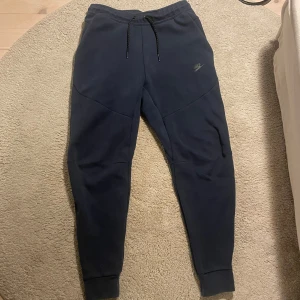 Svarta mjukisbyxor från Nike - Säljer ett par svarta mjukisbyxor från Nike med dragsko i midjan och ribbade muddar vid bensluten. Perfekta för en avslappnad stil eller träning. Byxorna har en diskret Nike-logga på sidan.