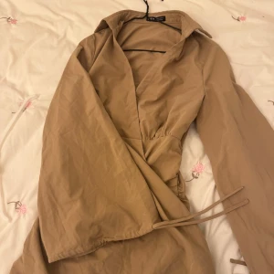 Beige omlottklänning - Säljer en stilren beige omlottklänning med långa ärmar och knytning i midjan. Perfekt för en elegant look. Klänningen har en klassisk krage och är tillverkad i ett mjukt material.