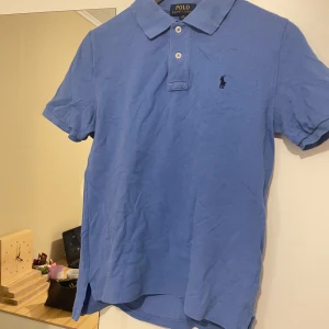 Blå pikétröja från Polo Ralph Lauren - Snygg blå pikétröja från Polo Ralph Lauren med klassisk krage och två knappar. Den har en liten broderad logga på bröstet. Perfekt för en stilren look.