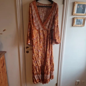 Blommig långklänning med volangdetaljer - Säljer en vacker långklänning i blommigt mönster med varma orange och lila toner. Klänningen har en omlott-design med v-ringning och knytband i midjan. Ärmarna är långa och vida med volangdetaljer. Perfekt för en bohemisk look!