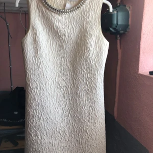 Champagnefärgad klänning med pärldetaljer - Elegant champagnefärgad klänning med pärlor runt halsringningen. Klänningen är ärmlös och har en strukturerad yta som ger en lyxig känsla. Perfekt för speciella tillfällen.