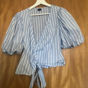 Randig omlottblus i blått och vitt - Säljer en söt randig omlottblus i blått och vitt med knytning i midjan. Blusen har puffiga trekvartsärmar och en v-ringad design. Perfekt för en somrig look!