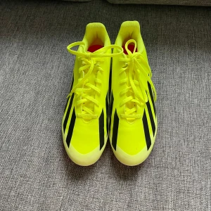 Gula fotbollsskor från Adidas - Säljer ett par gula fotbollsskor från Adidas med svarta detaljer och snörning. Skorna har en mönstrad sula med röda och vita dobbar, perfekt för snabba rörelser på planen.