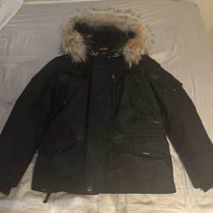 Svart parajumper jacka - perfekt nu till vintern! - Säljer en svart parkajacka från Parajumpers med pälsdetaljer på huvan. Jackan har flera praktiska fickor och en dragkedja framtill. Den är quiltad på insidan för extra värme och har en snygg orange detalj på baksidan.