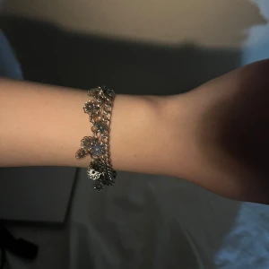 armband med blommor - Charmigt guldarmband med små blommor som hänger längs kedjan. Perfekt för att ge en elegant touch till din outfit. Armbandet har en fin glans och är lätt att matcha med andra smycken.