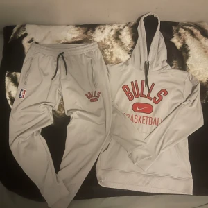 Grå hoodie från Nike med Bulls-tryck och byxor hel tracksuit  - Säljer en grå hoodie från Nike med Bulls-tryck och NBA-logga. Tröjan har en stor ficka fram och justerbar huva med svarta snören. Perfekt för basketfans! 🏀