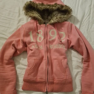 Rosa hoodie med päls från Abercrombie - Säljer en mysig rosa hoodie från Abercrombie med pälsfodrad huva och dragkedja. Den har tryck med siffrorna '1892' och 'Abercrombie' på framsidan. Perfekt för kyliga dagar! Står storlek M men skulle uppskatta till S! Ni kan alltid försöka pruta, det värsta är att ni kan få nej🩷